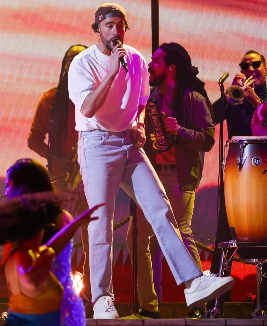 Bad Bunny performs on stage with his band, wearing a white t-shirt, jeans, and a baseball cap, singing into a microphone.