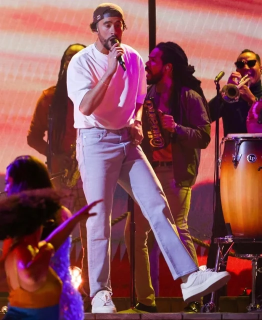 Bad Bunny performs on stage with his band, wearing a white t-shirt, jeans, and a baseball cap, singing into a microphone.