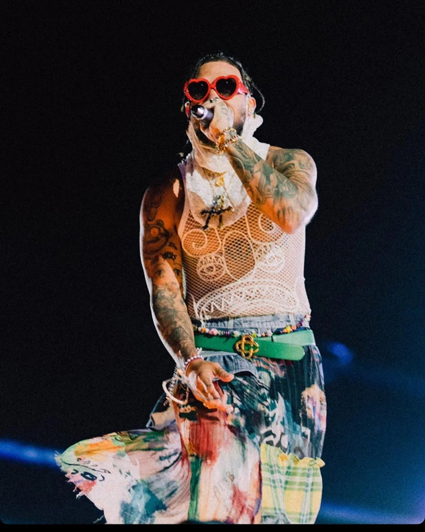 Singer performing on stage in heart-shaped sunglasses and mesh top. The concert setting is dark with blue lighting. Musician holds a microphone.