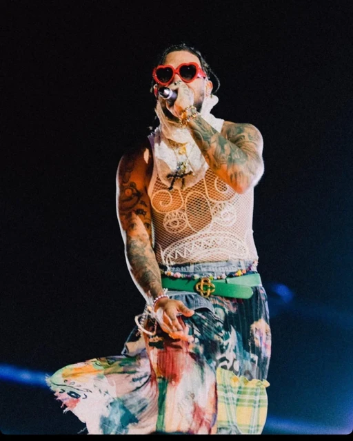 Singer performing on stage in heart-shaped sunglasses and mesh top. The concert setting is dark with blue lighting. Musician holds a microphone.