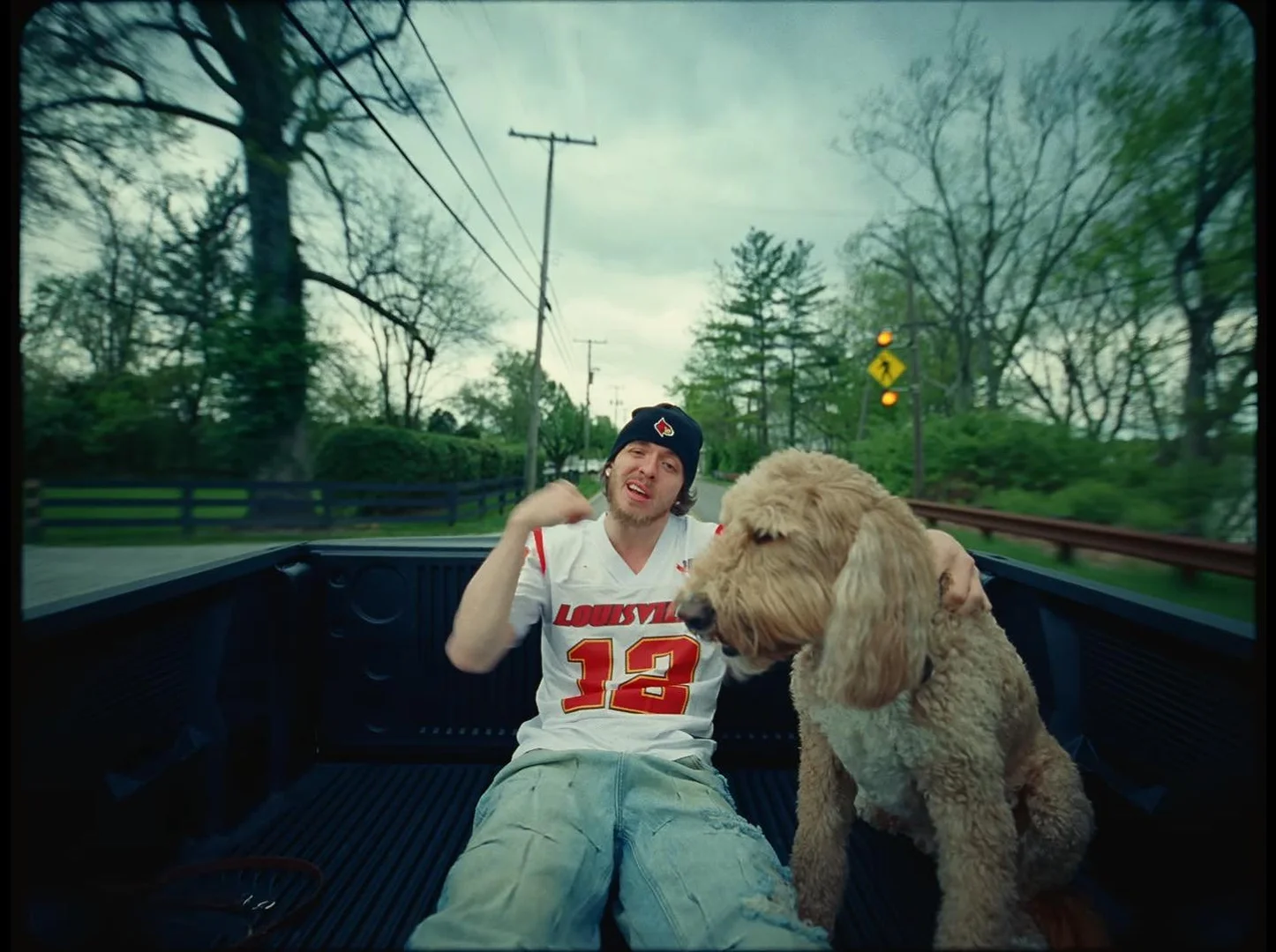 An adult male wearing a jersey and a beanie sits in the back of a truck with a golden doodle dog in a rural outdoor setting.