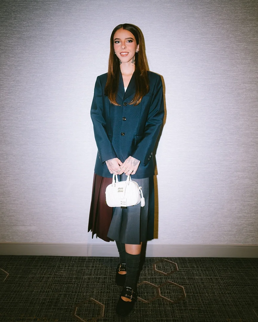 Full-shot of a woman with long brown hair posing in a navy blazer, pleated skirt, Mary Jane shoes, knee socks, and carrying a purse indoors.