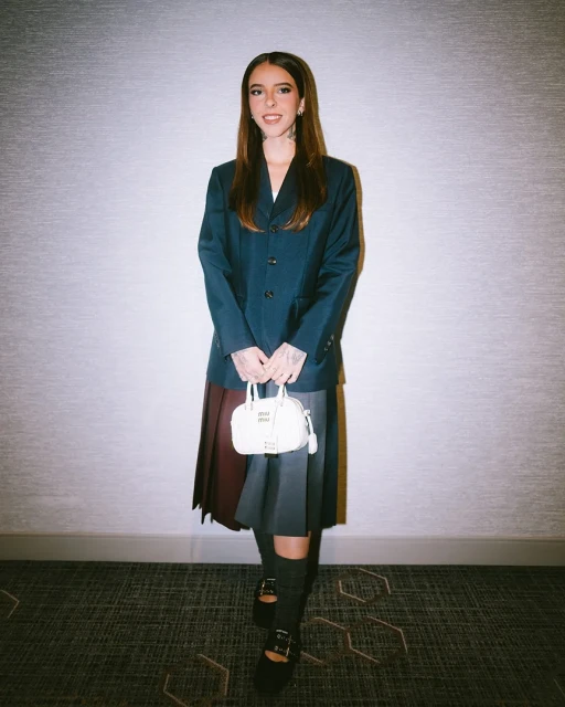 Full-shot of a woman with long brown hair posing in a navy blazer, pleated skirt, Mary Jane shoes, knee socks, and carrying a purse indoors.