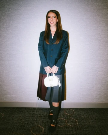 Full-shot of a woman with long brown hair posing in a navy blazer, pleated skirt, Mary Jane shoes, knee socks, and carrying a purse indoors.