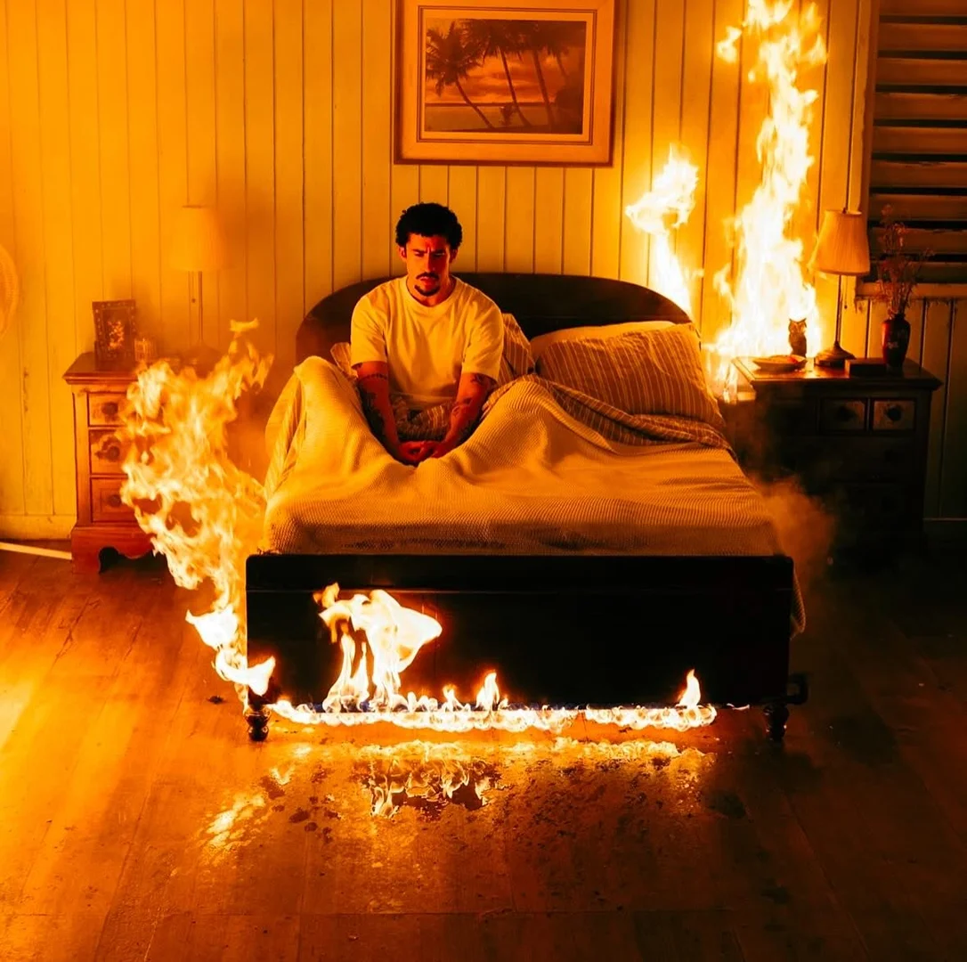 A man sits on a bed surrounded by flames in a surreal indoor setting with a framed painting on the wall, capturing an intense scene.