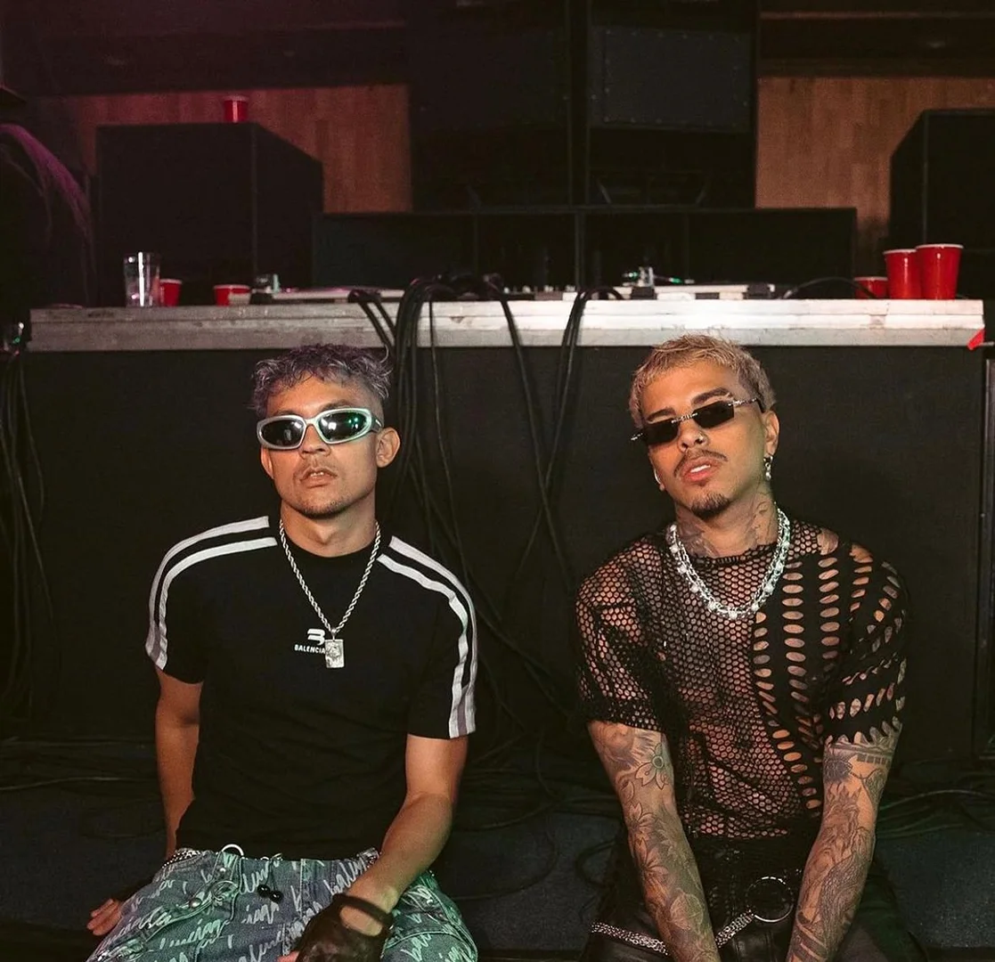 Two adult males pose in an indoor setting, showcasing streetwear fashion with accessories and tattoos in a medium shot with ambient lighting.