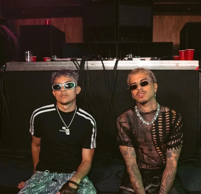 Two adult males pose in an indoor setting, showcasing streetwear fashion with accessories and tattoos in a medium shot with ambient lighting.