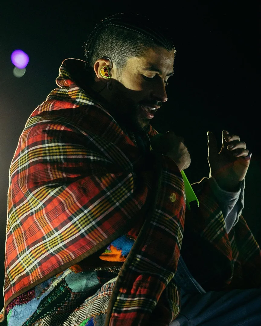 Adult male with cornrow braids wrapped in a plaid blanket, partially visible patterned shawl, lit by a bright spot on a dark stage.