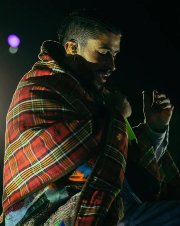 Adult male with cornrow braids wrapped in a plaid blanket, partially visible patterned shawl, lit by a bright spot on a dark stage.