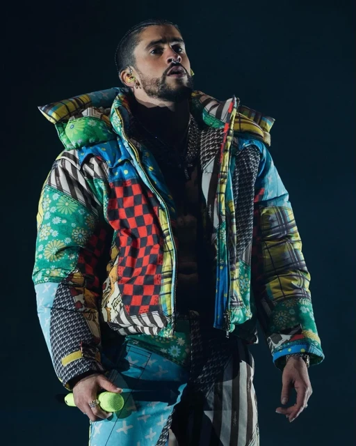 Bad Bunny performing on stage wearing a patterned puffer jacket and pants with a microphone in hand in a dark, moody atmosphere.