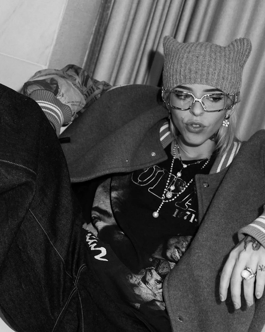 Black and white studio portrait of a young adult woman with a knit cap, glasses, jewelry, jacket, and dark clothing, sitting indoors.