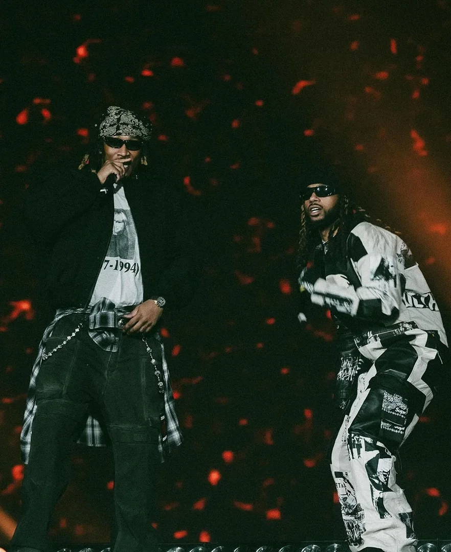 Two male hip-hop artists are performing on stage under warm lighting with one wearing a bandana and the other in patterned pants.