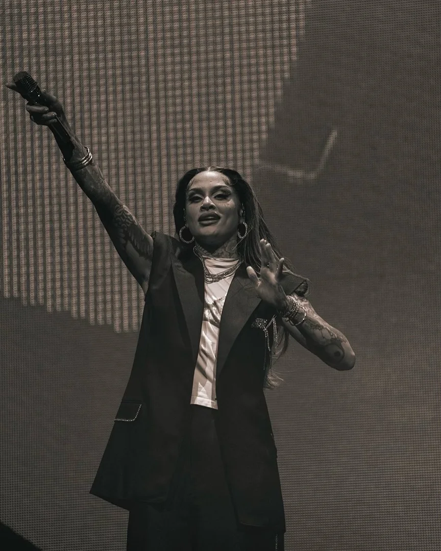 Monochrome shot of a woman singing on stage with arm raised, wearing a black sleeveless blazer, hoop earrings, and multiple bracelets.