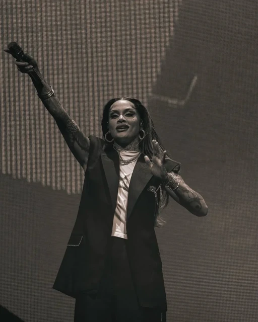 Monochrome shot of a woman singing on stage with arm raised, wearing a black sleeveless blazer, hoop earrings, and multiple bracelets.
