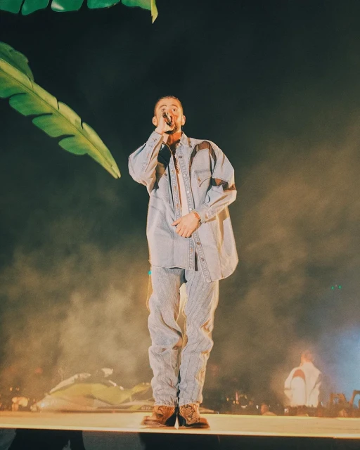 Singer Bad Bunny performs on stage in a light blue denim outfit under bright stage lights with green leaf decorations.