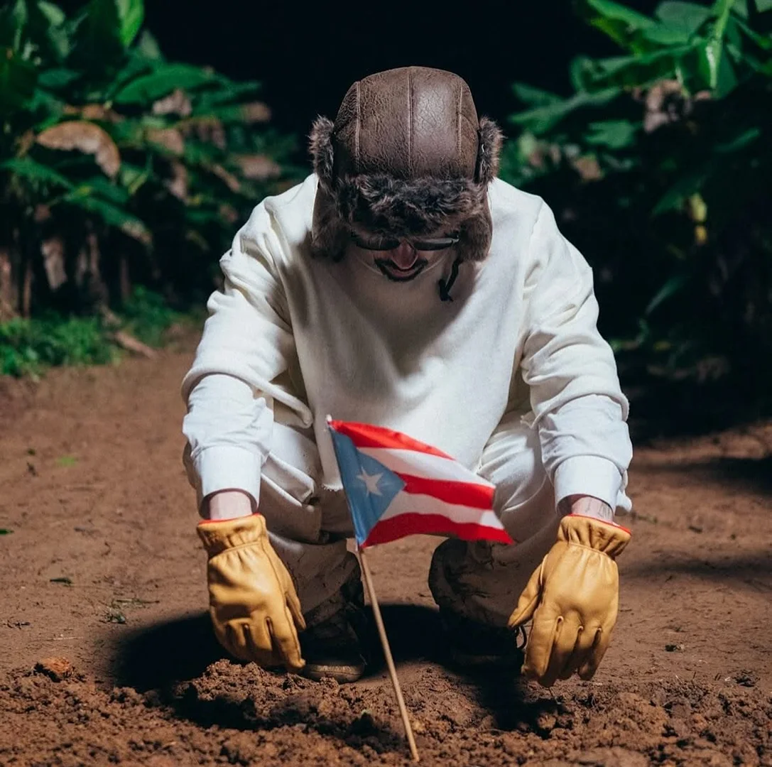 Person in earflap hat kneeling on dirt ground holding Puerto Rican flag, wearing gloves and a white sweatshirt, in a dark outdoor environment.