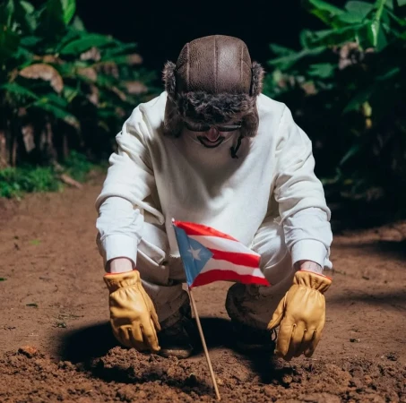 Person in earflap hat kneeling on dirt ground holding Puerto Rican flag, wearing gloves and a white sweatshirt, in a dark outdoor environment.