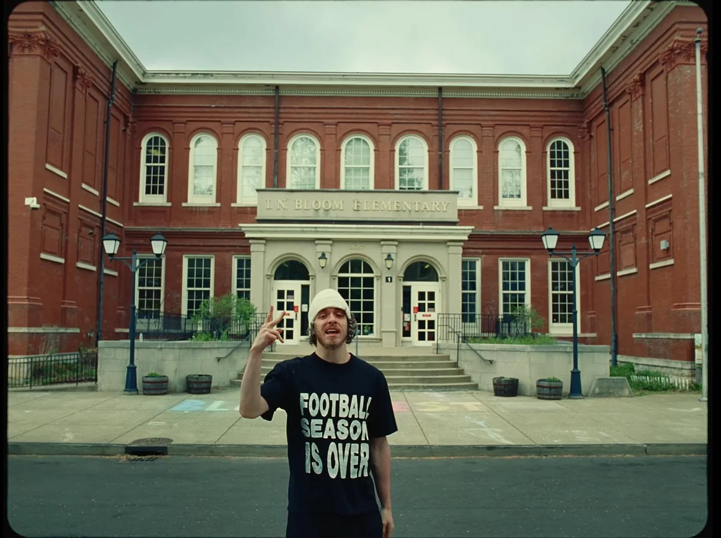Adult male wearing a beanie and t-shirt stands in front of a brick elementary school on a cloudy day, making a hand gesture.