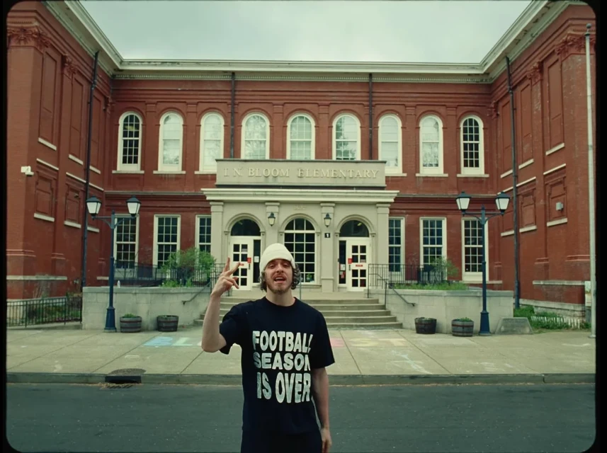 Adult male wearing a beanie and t-shirt stands in front of a brick elementary school on a cloudy day, making a hand gesture.