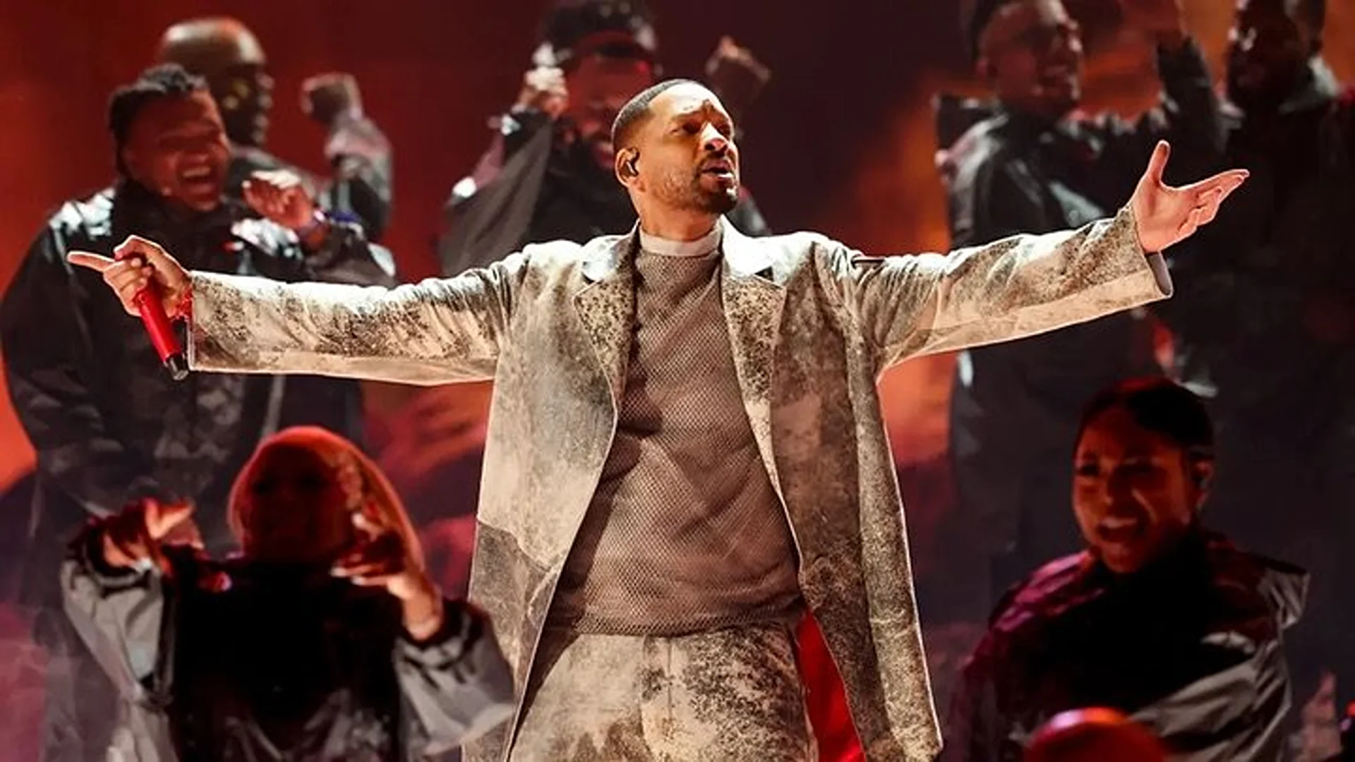 Singer on stage with outstretched arms, performing live to a backing choir. Warm stage lighting and a diverse group add to the atmosphere.