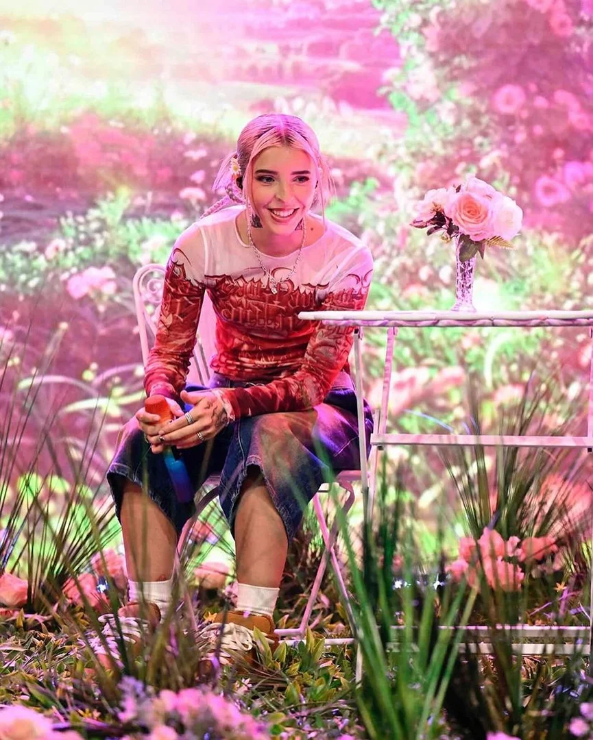A young woman with blonde hair sits at a white table in a field of flowers, smiling, and holding an apple. Roses are in a vase on the table.