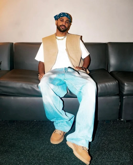 Candid shot of a seated adult male wearing blue tie-dye jeans, a white t-shirt, tan vest, and bandana in a neutral indoor setting.