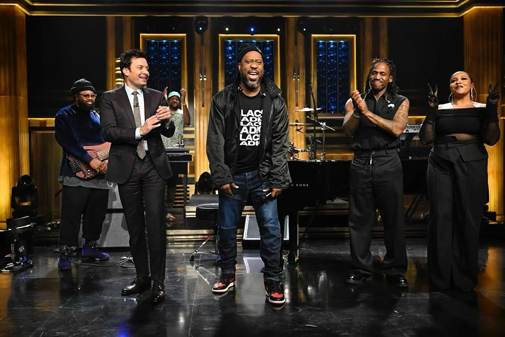 On stage, Jimmy Fallon and the Roots perform for The Tonight Show. Questlove stands near a piano, with Black Thought in the center.