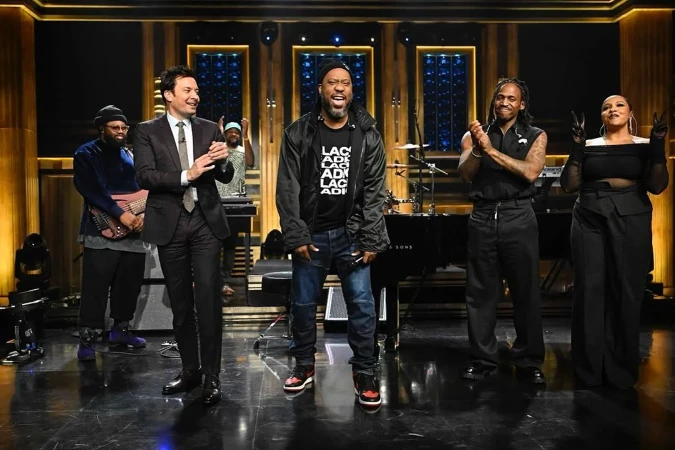 On stage, Jimmy Fallon and the Roots perform for The Tonight Show. Questlove stands near a piano, with Black Thought in the center.