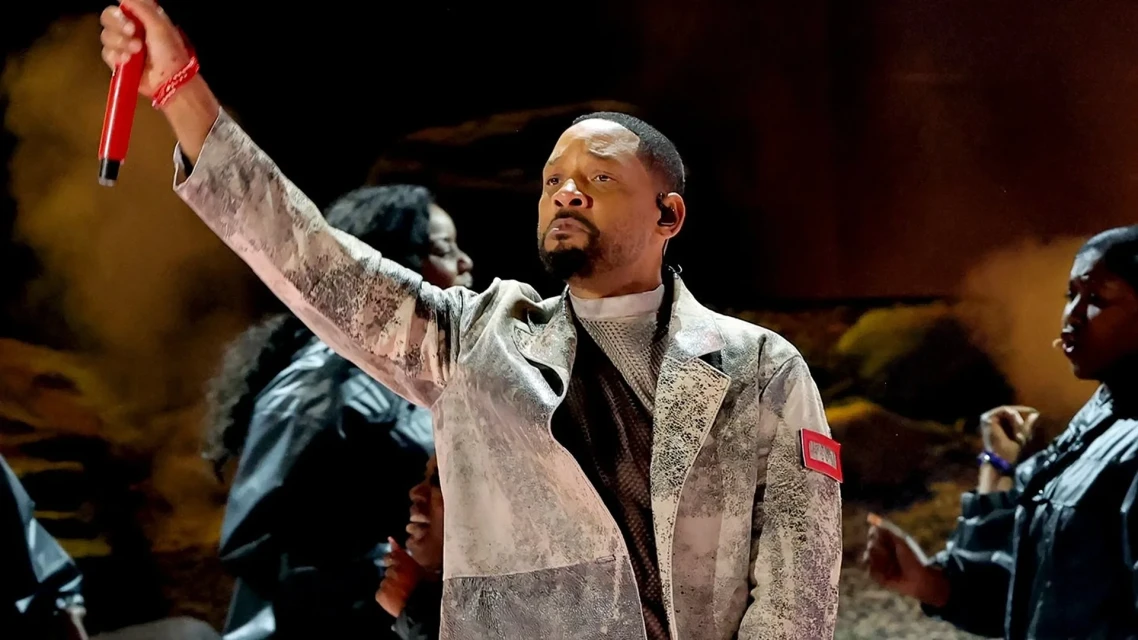 On stage, a distinguished adult male entertainer performs with a red microphone, wearing a textured white jacket amid background performers.