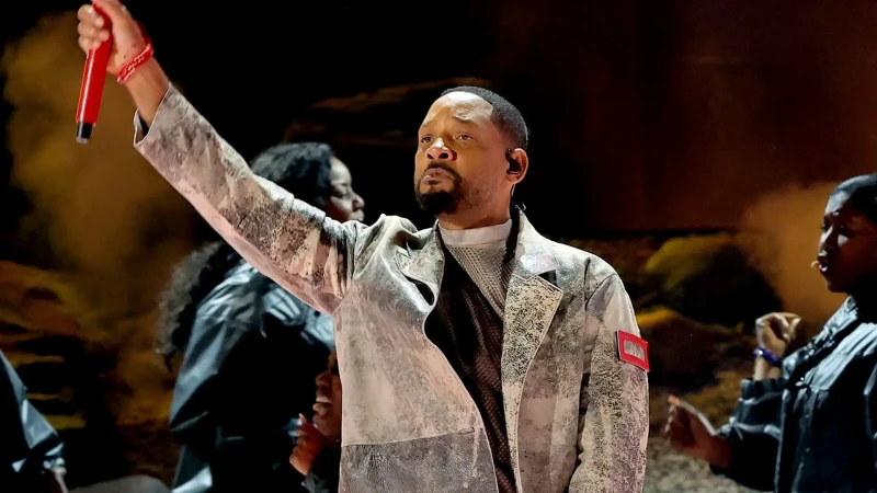 On stage, a distinguished adult male entertainer performs with a red microphone, wearing a textured white jacket amid background performers.