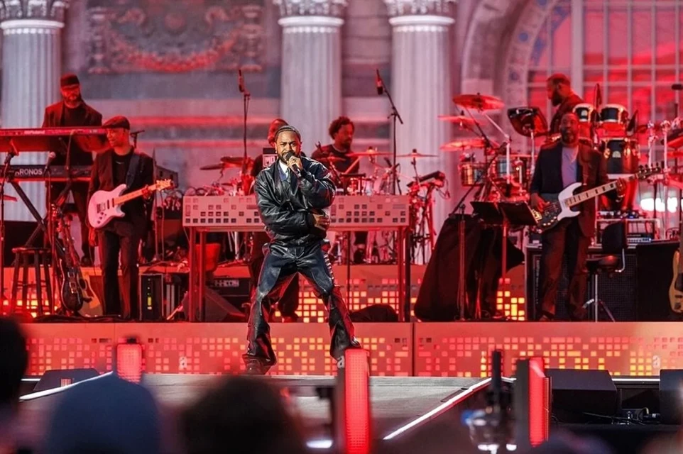 Musical performance on stage features a man in a dark outfit singing into a microphone, backed by a band with red lighting effects.