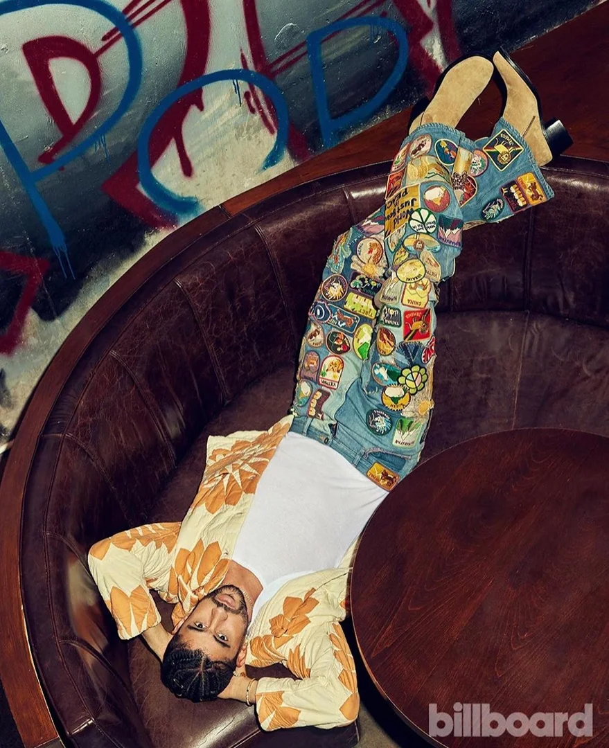 Singer Bad Bunny lounges in a leather chair with floral shirt, patch-covered jeans, and tan boots next to round table. Billboard logo visible.