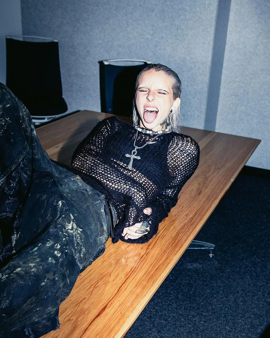A person with wet hair and a cross necklace lies on a wooden table, sticking out their tongue, with a gray wall in the background.