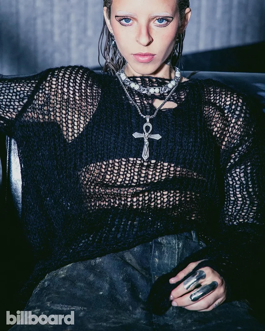 Fashion portrait of a fair-skinned person with white eyebrows and black eye makeup wearing a black mesh top and jewelry, posing indoors.