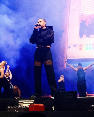 A man sings on stage in black hoodie and pants, backed by dancers and a blue screen during a concert performance with stage lighting.