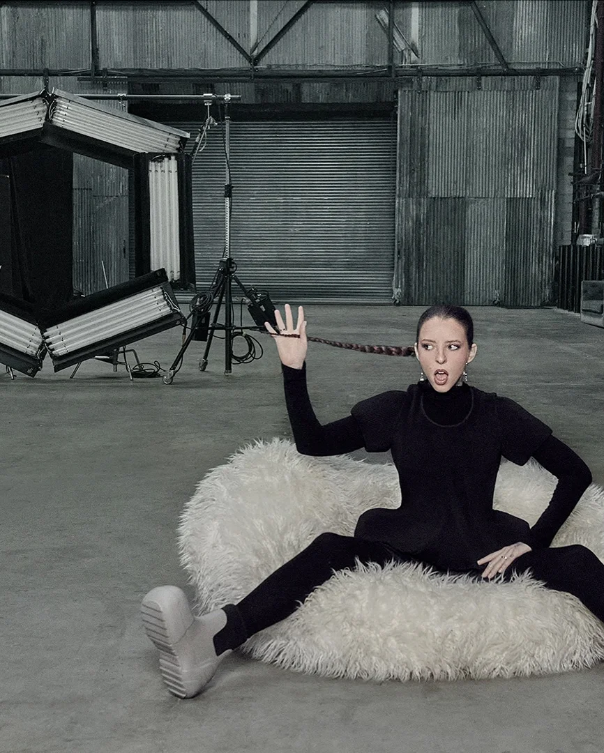 Woman in a black outfit posing on a white furry cushion in a photo studio, showcasing her long braid and wearing platform boots.