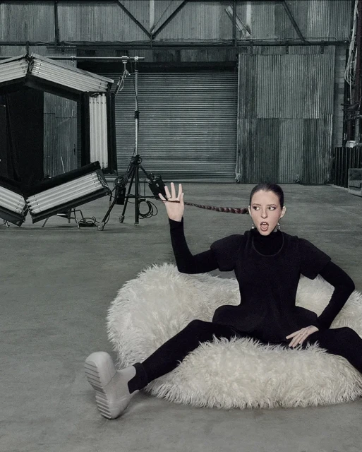 Woman in a black outfit posing on a white furry cushion in a photo studio, showcasing her long braid and wearing platform boots.