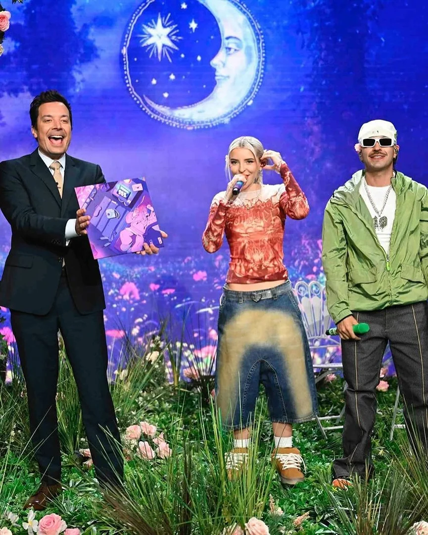 Three individuals pose on a set with grass and flowers, with a moon backdrop. One holds an album cover, others hold microphones.