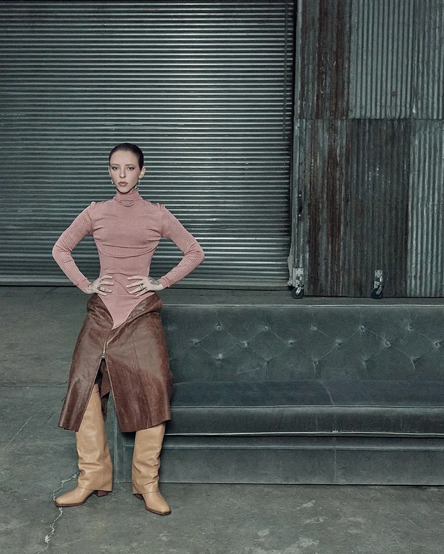 Woman poses in a pink turtleneck bodysuit, brown leather skirt, and beige boots against a metal industrial backdrop on a grey concrete floor.