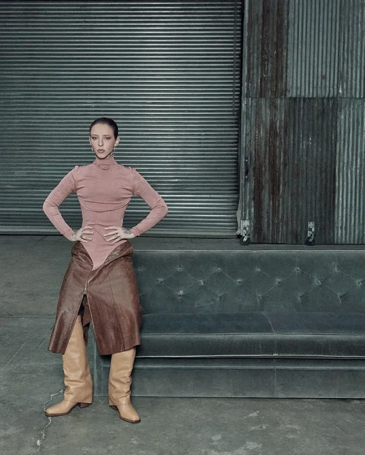 Woman poses in a pink turtleneck bodysuit, brown leather skirt, and beige boots against a metal industrial backdrop on a grey concrete floor.