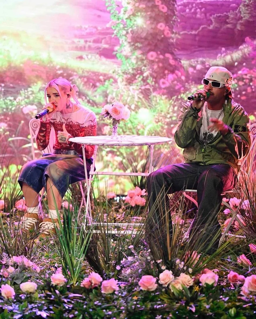 Two singers performing on a stage decorated with flowers and plants, with pink and green lighting and a floral backdrop.