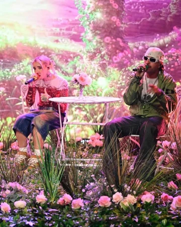 Two singers performing on a stage decorated with flowers and plants, with pink and green lighting and a floral backdrop.