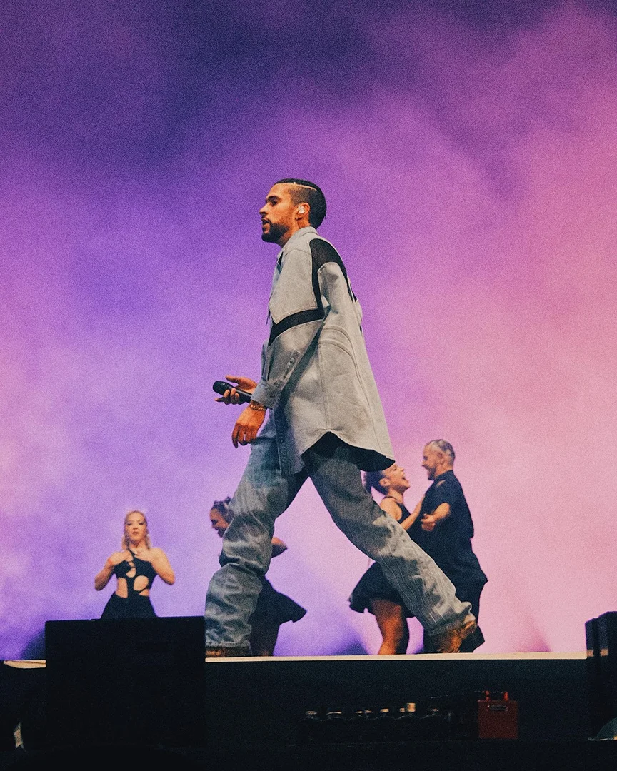 Singer Bad Bunny performing live on stage with backup dancers in a purple and pink-lit arena, wearing denim and holding a microphone.