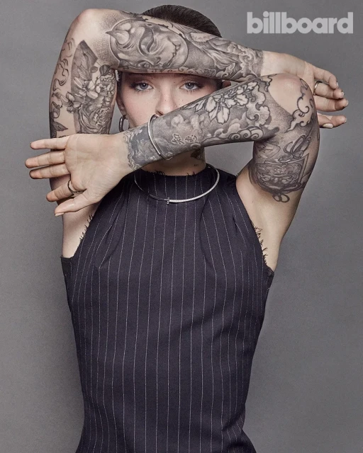 Portrait of a young woman with tattoos covering her arms, posing with her arms crossed over her face, wearing a pinstripe top.