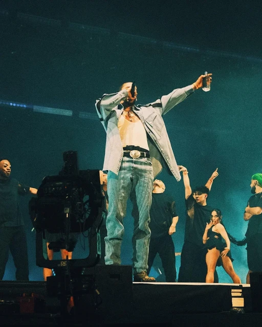 A singer performs on stage holding a water bottle, wearing a denim jacket, jeans, and a white tank top, backed by dancers.
