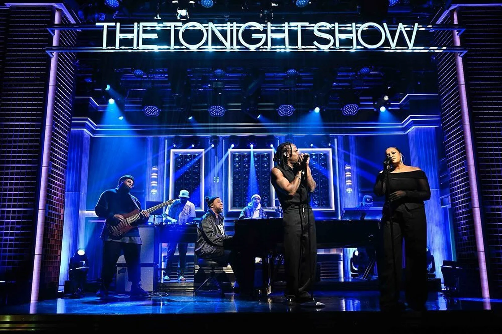 Musical performance on The Tonight Show stage featuring a diverse group of musicians against a backdrop of blue lighting and architectural details.