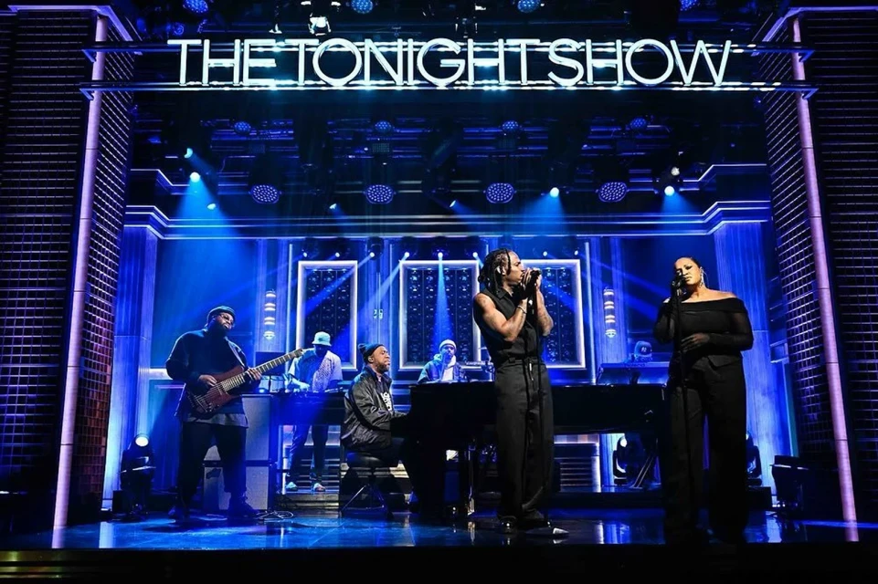 Musical performance on The Tonight Show stage featuring a diverse group of musicians against a backdrop of blue lighting and architectural details.