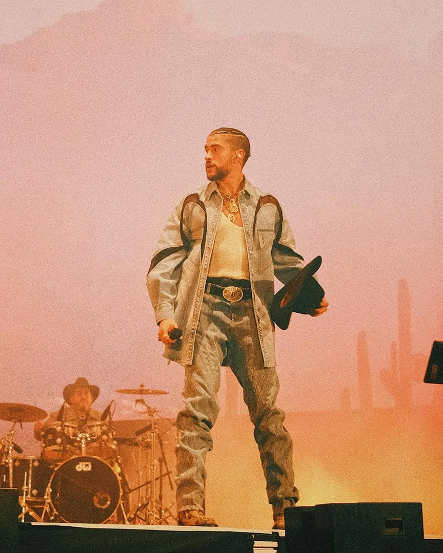 Bad Bunny performing on stage with a cowboy hat and denim outfit, a drummer is visible in the background.