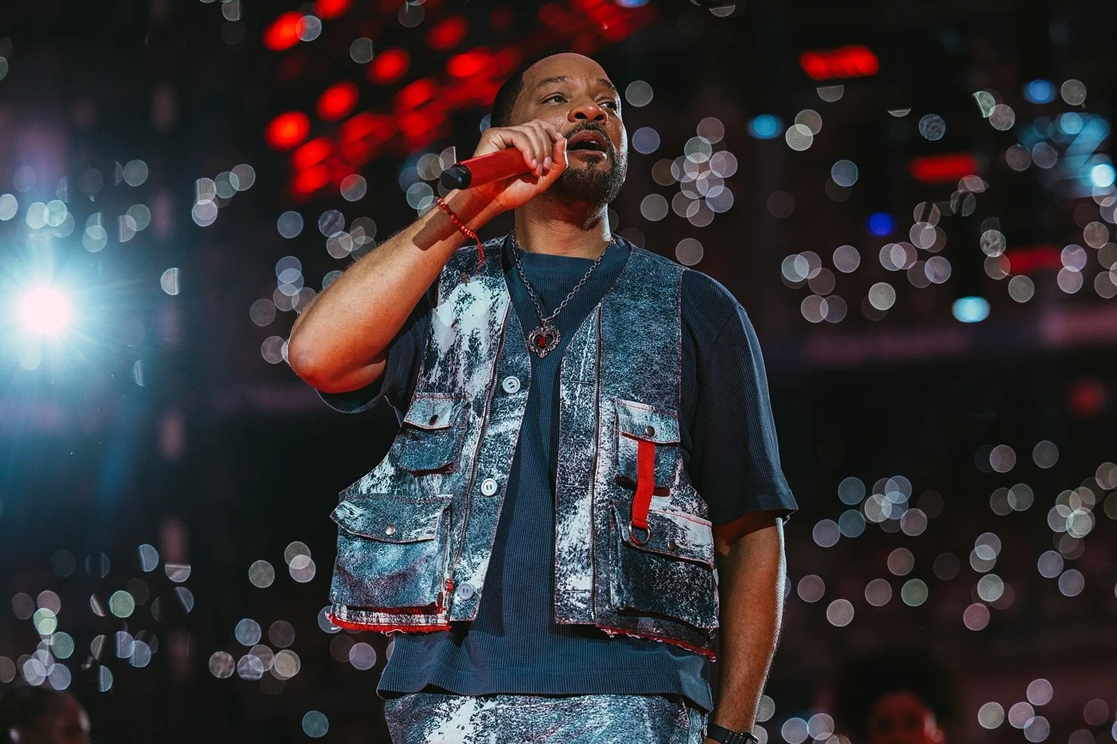 Will Smith performs on stage, singing into a red microphone while wearing a vest, at an indoor music venue filled with lights.
