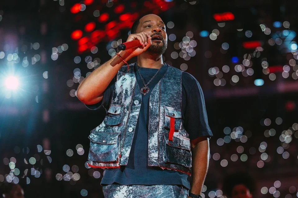 Will Smith performs on stage, singing into a red microphone while wearing a vest, at an indoor music venue filled with lights.